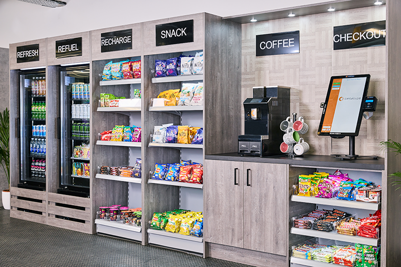 A medium sized light grey wood micro market with 2 coolers, a coffee machine and large self-checkout kiosk.